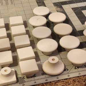 May include: An assortment of white cabinet knobs and pulls in various shapes, including square and round designs. The knobs are arranged on a grid-patterned surface, showcasing their different sizes and styles. A ruler is visible along the edge.