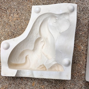 May include: Two white plaster molds, likely for casting sculptures, are shown. One mold features a detailed relief of a reclining figure, while the other is partially visible. The molds have rounded edges and are set against a textured, light-colored surface.