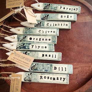 May include: A collection of ceramic garden markers with names of herbs like parsley, sage, and rosemary. Each marker is rectangular with a pointed end, featuring a speckled blue-green design and black lettering. The markers are arranged on a wooden tray.