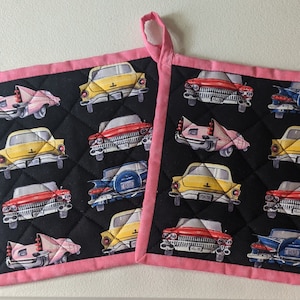 May include: Two pink and black quilted hot pads with a vintage car pattern. The cars are yellow, red, blue, and pink. The hot pads have a pink border.