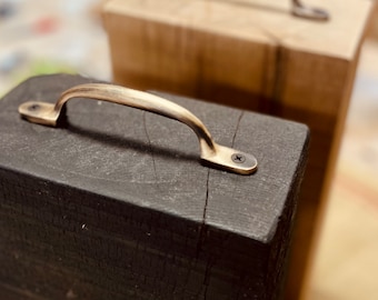 May include: Two rectangular wooden blocks, one finished in matte black and the other in a natural light brown wood tone. Each block features a curved, metallic handle in a brushed brass finish, secured to the top surface with two small screws.