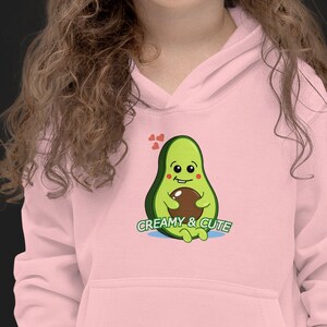 Può includere: Una felpa con cappuccio rosa con un avocado disegnato che tiene un avocado più piccolo. L'avocado ha un viso sorridente e il testo "Creamy & Cute" è stampato sotto.