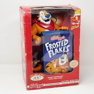 May include: A red, white, and blue box with a picture of Tony the Tiger on it. The box says "Kellogg's Frosted Flakes" and "Anniversary Collection". The box is for a battery-operated talking Tony the Tiger bank.