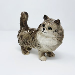 Puede incluir: Una figurita de cerámica de un gato marrón y blanco con pelo largo. El gato está de pie sobre sus cuatro patas y tiene la cola enroscada detrás de él.