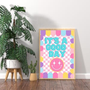 Peut inclure: Une impression encadrée avec un fond à carreaux rose, jaune et bleu. L'impression présente le texte "It's a good day" en police bleue avec un smiley rose avec deux étoiles en dessous.