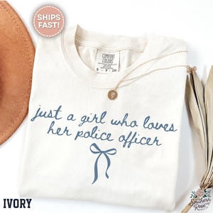 May include: Ivory-colored t-shirt with the text "just a girl who loves her police officer" in blue script. A blue bow graphic is below the text. A gold necklace with a pendant is on the shirt.