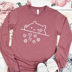 May include: A long-sleeved, maroon t-shirt with a white graphic of a cat peeking over a line of dice. The cat has a cute expression and is surrounded by dice.