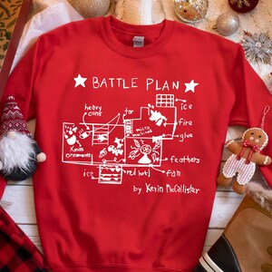 May include: Red Christmas sweater with a white graphic of a house plan labeled "Battle Plan" with various items like "heavy cans", "ice", "fire", "glue", "feathers", "fan", and "Christmas ornaments". The text "by Kevin McCallister" is at the bottom of the plan.
