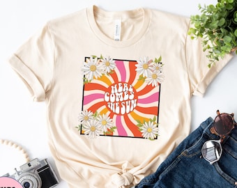 May include: A beige t-shirt with a retro-style graphic featuring a sunburst design and the text "Here Comes The Sun" surrounded by white daisies.