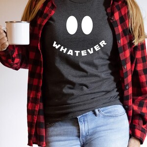 May include: A person wearing a gray t-shirt with a white smiley face and the text "WHATEVER" printed on it. The person is also wearing a red and black plaid shirt and blue jeans.