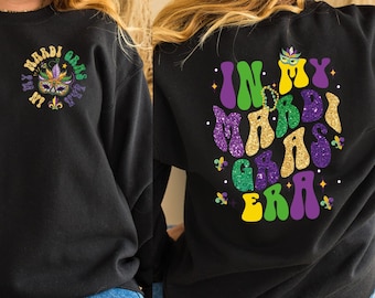 May include: Black sweatshirt with a green, gold, and purple glitter design that says "In My Mardi Gras Era". The front of the sweatshirt has a smaller design with a mask and the words "Mardi Gras I'm In My Era".