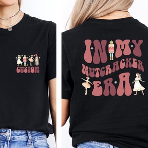 May include: Black t-shirt with a pink and white graphic on the front that says "CUSTOM" and a pink and white graphic on the back that says "IN MY NUTCRACKER ERA".