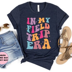 K&ouml;nnte beinhalten: Ein marineblaues T-Shirt mit dem Text "IN MY FIELD TRIP ERA" in bunter Retro-Schrift.