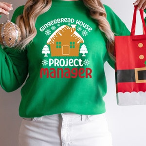 May include: A green sweatshirt with the text "Gingerbread House Project Manager" and a gingerbread house graphic. The person is holding a red and white gift bag with a Santa Claus design.