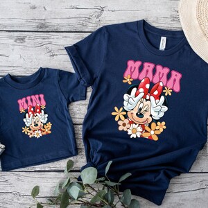 May include: Navy blue t-shirts with matching designs. One shirt says "MAMA" and the other says "MINI" in pink lettering. Both shirts feature a cartoon character with a red bow and flowers. A straw hat is in the background.