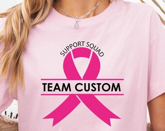 May include: A light pink t-shirt with the words "SUPPORT SQUAD" above a pink ribbon graphic and the words "TEAM CUSTOM" below. The shirt is a crew neck style.