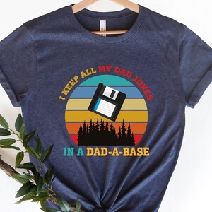 May include: A navy blue t-shirt with a retro sunset graphic and the text "I keep all my dad jokes in a dad-a-base". The graphic features a floppy disk flying through the air.
