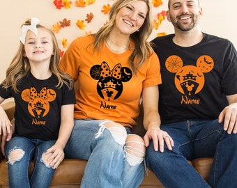 May include: A family of three wearing matching Halloween shirts. The shirts feature a black and white Mickey Mouse head with a cruise ship, bats, and a spider web. The shirts are orange and black. The text "Name" is on each shirt.