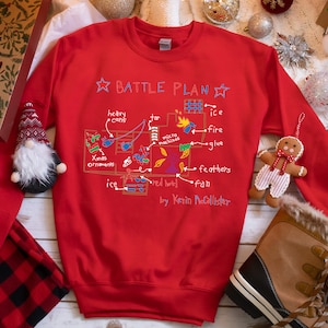May include: Red Christmas sweater with a white outline of a house and the words "Battle Plan" with a list of items to use for a Christmas battle. The items include heavy cans, ice, fire, glue, feathers, and a fan.