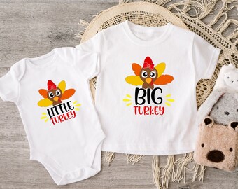 May include: Two white shirts with a Thanksgiving theme. One shirt says "Big Turkey" with a cartoon turkey wearing a bow. The other shirt says "Little Turkey" with a smaller cartoon turkey wearing a bow.