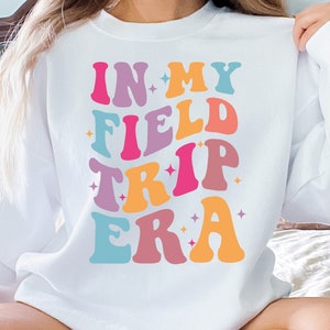 K&ouml;nnte beinhalten: Wei&szlig;er Pullover mit dem Text "IN MY FIELD TRIP ERA" in bunter Retro-Schrift.