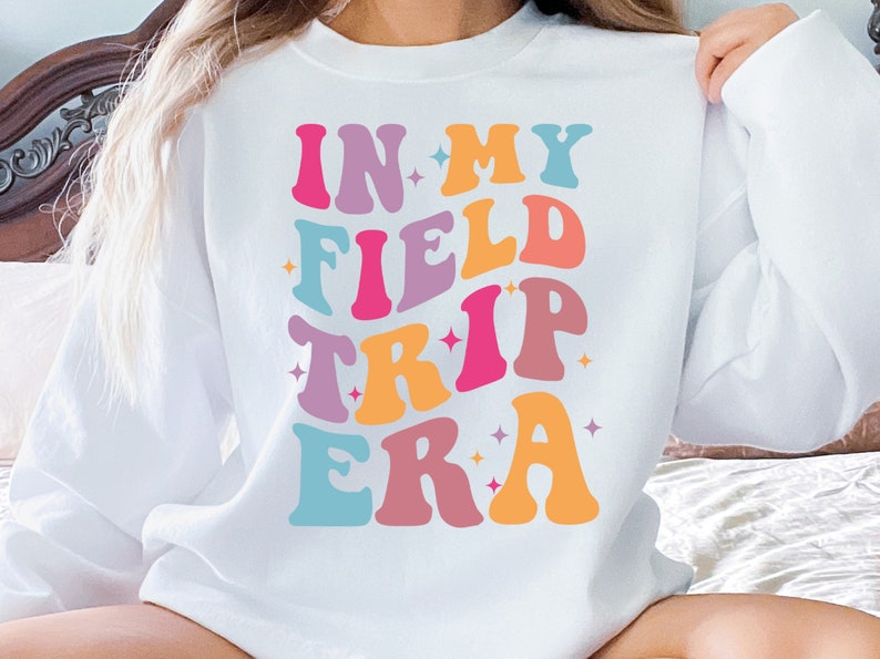 K&ouml;nnte beinhalten: Wei&szlig;er Pullover mit dem Text "IN MY FIELD TRIP ERA" in bunter Retro-Schrift.