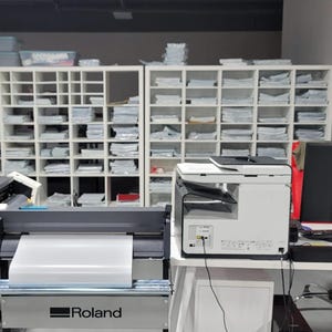 May include: A white office with a white printer, a black computer monitor, and a large white and gray plotter with the word "Roland" on it. There are white shelves filled with white paper on the left and right sides of the image.