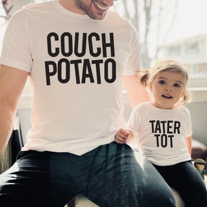 May include: A white t-shirt with the text "COUCH POTATO" in black lettering. A smaller white t-shirt with the text "TATER TOT" in black lettering is worn by a child.