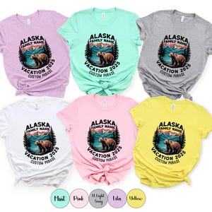Peut inclure: Plusieurs t-shirts de couleurs vari&eacute;es avec un motif circulaire repr&eacute;sentant un ours, des montagnes et des arbres. Le texte indique "Alaska Family Name Vacation 2025 Custom Phrase". Les t-shirts sont en menthe, rose, gris clair, lilas, jaune et blanc.