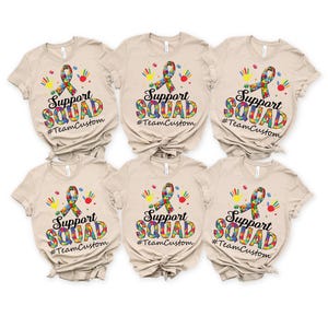 May include: Six beige t-shirts with a colorful puzzle piece ribbon, handprints, and the text "Support Squad #TeamCustom" printed on the front.