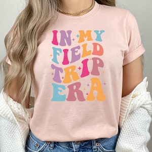 K&ouml;nnte beinhalten: Ein pinkfarbenes T-Shirt mit dem Text "In My Field Trip Era" in einer Retro-Schriftart mit bunten Buchstaben und Sternen.