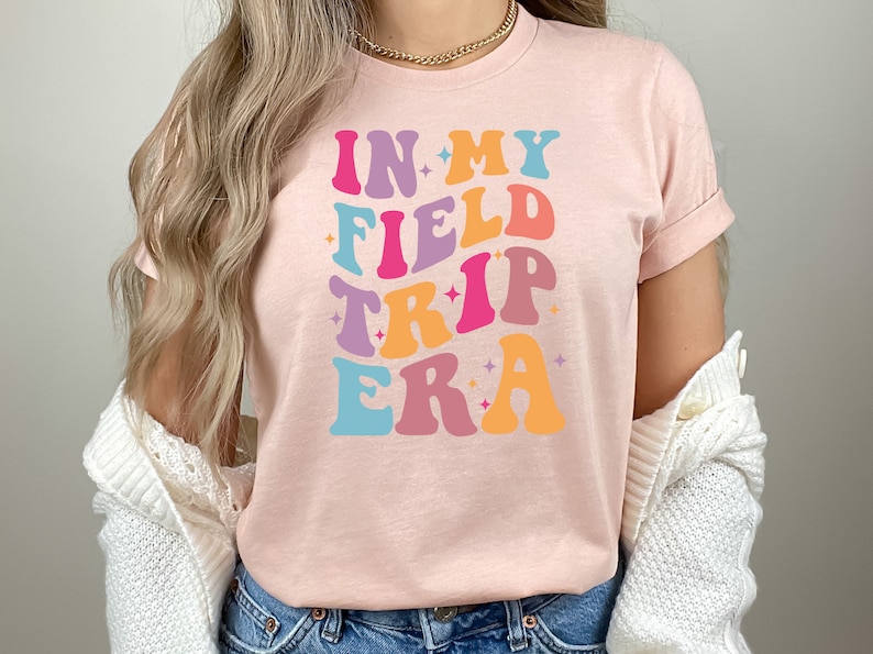 K&ouml;nnte beinhalten: Ein pinkfarbenes T-Shirt mit dem Text "In My Field Trip Era" in einer Retro-Schriftart mit bunten Buchstaben und Sternen.
