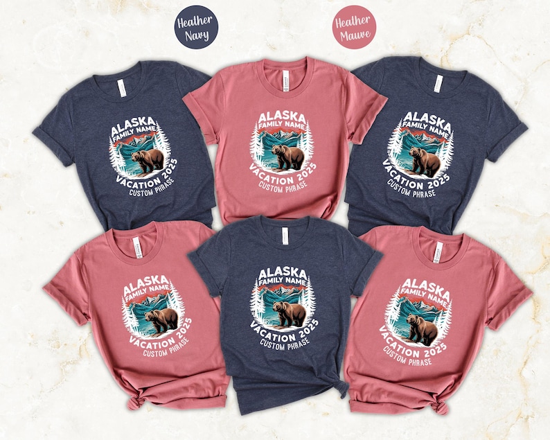 May include: A collection of short-sleeved t-shirts in heather navy and heather mauve. Each shirt features a circular graphic with a bear, mountains, and the text "ALASKA FAMILY NAME VACATION 2025 CUSTOM PHRASE."