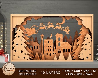 May include: A laser cut wooden Christmas scene with a town, trees, and Santa Claus flying in his sleigh with reindeer. The scene is framed in a wooden frame. The text "10 LAYERS" and "DIGITAL FILES FOR LASER CUT" is visible below the image.
