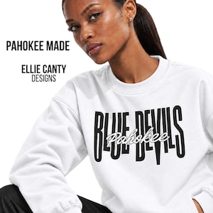 May include: A white crewneck sweatshirt featuring the black text BLUE DEVILS in a bold, vertical font with the word Pahokee written in a white script overlay. The text PAHOKEE MADE and ELLIE CANTY DESIGNS appears in black on the upper left side.