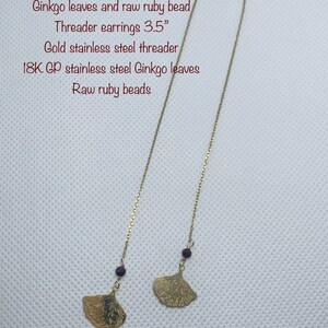 May include: A pair of gold-tone threader earrings featuring ginkgo leaf charms and small ruby beads. The earrings are 3.5 inches long and made with 18K gold plated stainless steel.