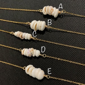May include: Five delicate necklaces with gold-colored chains. Each necklace features a different design of small, white seashells strung together. The seashells have natural variations in color and texture, creating a unique look.