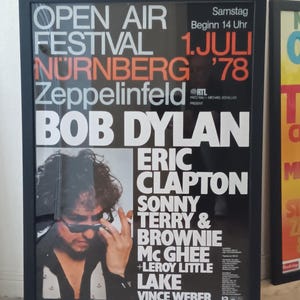 May include: Framed concert poster for the Open Air Festival in Nuremberg, Germany, in 1978, featuring Bob Dylan, Eric Clapton, and others. The poster has a black frame and a black background with white and orange text. A photo of Bob Dylan is included.