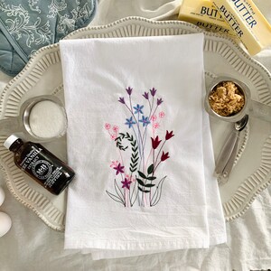 May include: A white kitchen towel with embroidered flowers in shades of pink, purple, blue, and green. The towel is on a white platter with a small bowl of brown sugar, a bottle of vanilla extract, and two white eggs.
