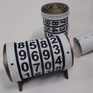 May include: Three-piece puzzle with a cylindrical design. The puzzle pieces are light gray with black numbers and dark brown accents. One piece is a cylinder with a lid, another is a cylinder with numbers, and the third is a tube with a stand.