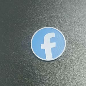 Pu&ograve; includere: Un logo di Facebook rotondo, azzurro e bianco. Il logo presenta una 'f' bianca su sfondo azzurro, circondata da un bordo bianco. Il logo &egrave; posto su una superficie grigio scuro.