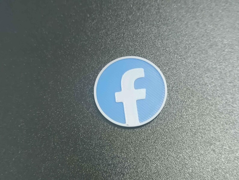 Pu&ograve; includere: Un logo di Facebook rotondo, azzurro e bianco. Il logo presenta una 'f' bianca su sfondo azzurro, circondata da un bordo bianco. Il logo &egrave; posto su una superficie grigio scuro.
