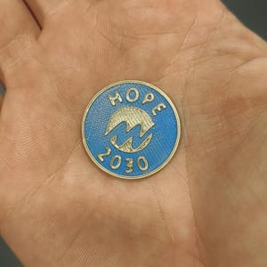 Pu&ograve; includere: Una moneta rotonda, blu e oro, con la scritta "HOPE" in alto e il numero "2030" in basso. Una "M" stilizzata al centro. La moneta &egrave; tenuta in una mano. Diametro: circa 2.5 cm.
