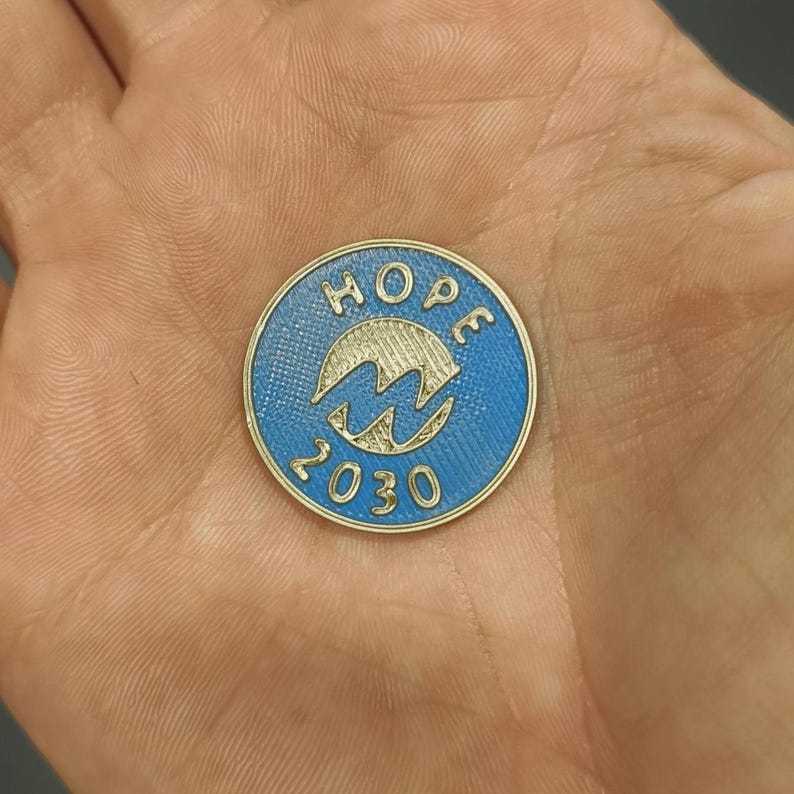 Pu&ograve; includere: Una moneta rotonda, blu e oro, con la scritta "HOPE" in alto e il numero "2030" in basso. Una "M" stilizzata al centro. La moneta &egrave; tenuta in una mano. Diametro: circa 2.5 cm.