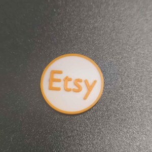 Pu&ograve; includere: Logo Etsy rotondo, bianco con scritta arancione e bordo abbinato. Il logo &egrave; posto su una superficie grigio scuro.