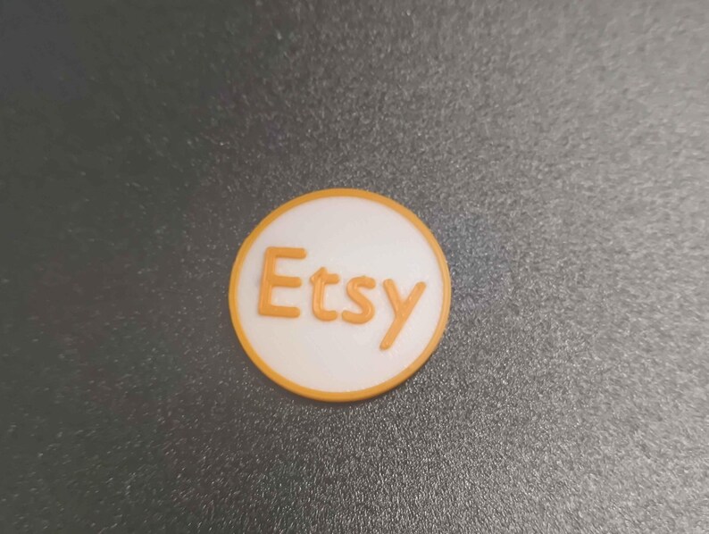 Pu&ograve; includere: Logo Etsy rotondo, bianco con scritta arancione e bordo abbinato. Il logo &egrave; posto su una superficie grigio scuro.
