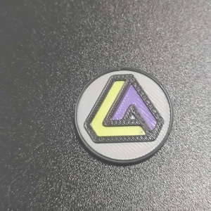 Pu&ograve; includere: Una moneta rotonda color argento con un design geometrico. Il design presenta un triangolo giallo e viola all'interno di un contorno nero, su uno sfondo argentato. La moneta &egrave; probabilmente un oggetto da collezione o un gettone.