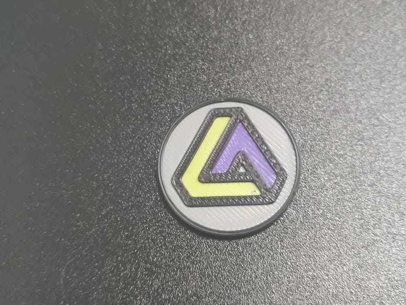 Pu&ograve; includere: Una moneta rotonda color argento con un design geometrico. Il design presenta un triangolo giallo e viola all'interno di un contorno nero, su uno sfondo argentato. La moneta &egrave; probabilmente un oggetto da collezione o un gettone.