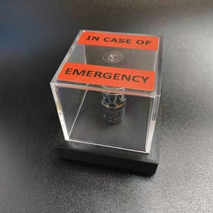 Puede incluir: Un pequeño objeto metálico encerrado en una caja de acrílico transparente con una base negra. La caja tiene una etiqueta roja que dice "IN CASE OF EMERGENCY" en texto blanco. El objeto está centrado dentro de la caja.