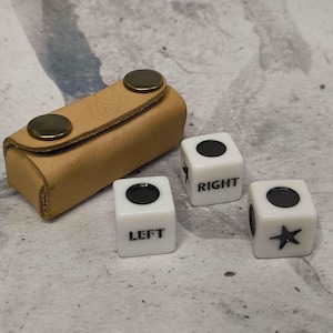 May include: A tan leather dice case with two brass-colored snap closures. Three white dice with black markings are next to the case. Two dice have a black dot, one has a star, and the other says "LEFT" and "RIGHT".